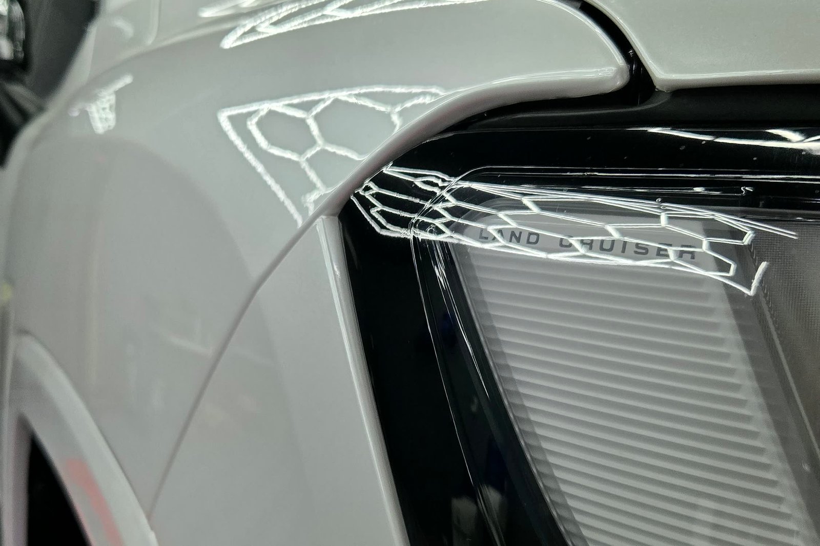 Close view of the front fender meeting the headlight on a white Toyota Land Cruiser 300, the paint protection film edge tucked into the panel gap and the ceiling lights reflecting unbroken across the panel